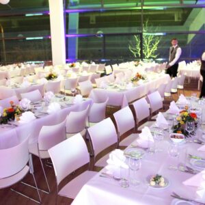 Bildquelle: Stadion Dresden Projektgesellschaft mbH & Co. KG Ein großer, elegant gedeckter Veranstaltungsraum im Rudolf-Harbig-Stadion in Dresden, vorbereitet für ein formelles Abendessen oder eine Feier. Zahlreiche lange Tische sind mit weißen Tischdecken, weißem Geschirr und Gläsern gedeckt und mit farbenfrohen Blumenarrangements dekoriert. Weiße Stühle stehen an den Tischen. Im Hintergrund sind zwei Kellner in Westen und weißen Hemden zu sehen. Die großen Fenster im Hintergrund lassen den Blick nach draußen zu.