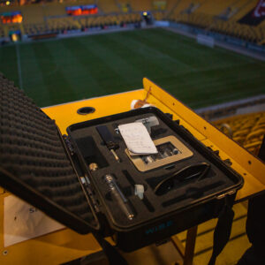 Ein offener, schwarzer Transportkoffer steht auf einem gelben Geländer, wahrscheinlich auf einer Tribüne in einem Stadion. Im Koffer sind verschiedene technische Geräte und Zubehörteile ordentlich in Schaumstoff-Aussparungen angeordnet, darunter Kabel, ein Messgerät und andere kleine Utensilien. Im Hintergrund ist unscharf ein grünes Fußballfeld und die gelben Sitzreihen eines Stadions zu erkennen, was darauf hindeutet, dass die Szene im Dynamo-Dresden-Stadion spielt. Die Beleuchtung lässt vermuten, dass es entweder Dämmerung ist oder die künstliche Beleuchtung des Stadions eingeschaltet ist.