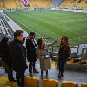 Eine Gruppe von vier Personen, drei Männer und eine Frau, stehen auf einer gelben Tribüne in einem Fußballstadion und blicken auf das Spielfeld. Die Frau in der Mitte, die eine graue Jacke trägt und ein Tablet in der Hand hält, zeigt mit dem Finger in die Ferne, als würde sie etwas erklären. Die anderen Personen schauen aufmerksam zu. Die leeren gelben Sitzreihen des Stadions erstrecken sich um das grüne Spielfeld herum. Im Hintergrund sind auf der Tribüne Buchstaben zu erkennen, die das Wort "DRESDEN" bilden.