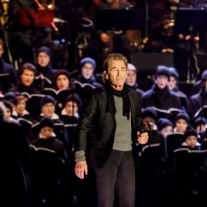 Foto: © 2018 Claudia Jacquemin Ein mittlerer Schuss eines Mannes, der auf einer Bühne steht und in ein Mikrofon singt. Er trägt einen dunklen Anzug mit einem schwarzen Schal und hat kurzes, gelocktes, blondes Haar. Im Hintergrund befindet sich ein Chor von Menschen, die in dunklen Mänteln und Mützen gekleidet sind. Ein Teil der Schulter eines anderen Mannes ist im Vordergrund links sichtbar.