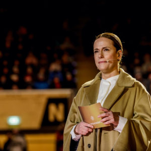 Foto©: Claudia Jacquemin Eine Frau mittleren Alters, die einen hellbraunen Mantel über einer weißen Bluse trägt, steht auf einer Bühne und hält ein goldenes Buch. Sie hat ein Mikrofon an ihrem Kragen befestigt. Im Hintergrund ist ein undeutliches Publikum und ein großes "N" zu sehen, das auf das Rudolf-Harbig-Stadion in Dresden hinweist.