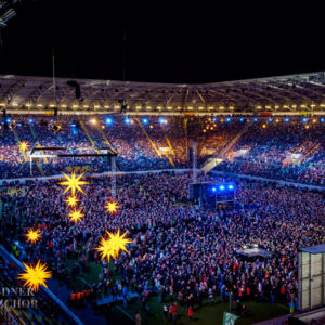 Foto: © 2018 Claudia Jacquemin Anfrage erfolgreich Ein hochauflösendes Luftbild vom Inneren des Rudolf-Harbig-Stadions in Dresden bei Nacht, gefüllt mit einer großen Menschenmenge, die eine Live-Musikveranstaltung besucht. Die Bühne ist am Ende des Stadions aufgebaut und ist von hellen Lichtern und Effekten beleuchtet. Mehrere leuchtende, sternförmige Dekorationen hängen über der Menge. Der Himmel ist dunkel, aber das gesamte Stadion ist gut beleuchtet. In der unteren linken Ecke des Bildes steht "DRESDNER KREUZCHOR".