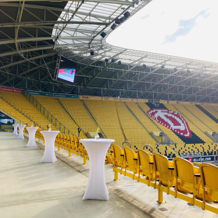 Das Bild zeigt einen Teil des Rudolf-Harbig-Stadions in Dresden, der offenbar für eine Veranstaltung vorbereitet ist. Im Vordergrund sind mehrere mit weißen Tüchern überzogene Stehtische auf einem gepflasterten Bereich zu sehen, der sich direkt vor den Tribünen befindet. Rechts davon sind gelbe Stadionsitze zu erkennen, die sich in Reihen nach oben erstrecken. Die Sitze bilden auch ein großes, weißes Logo, das stark an das Wappen von Dynamo Dresden erinnert. Im Hintergrund erstreckt sich die beeindruckende Dachkonstruktion des Stadions, die teilweise durchsichtig ist und den Blick auf den Himmel freigibt. Ein großer Videowürfel hängt unter dem Dach. Die Atmosphäre ist hell und offen, was auf einen sonnigen Tag hindeutet. Das Bild vermittelt den Eindruck einer exklusiven oder VIP-Area, die den Besuchern einen direkten Blick auf das Spielfeld und eine komfortable Umgebung bietet.