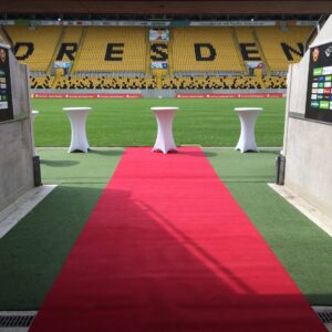 Das Bild zeigt einen Ausgang aus einem Stadiongebäude, der auf das Spielfeld führt. Ein leuchtend roter Teppich ist über einen grünen Kunstrasen ausgerollt und erstreckt sich von einem Tunnel oder Gang bis auf das eigentliche Spielfeld. Neben dem roten Teppich stehen drei weiße Stehtische auf dem grünen Kunstrasen. Im Hintergrund sind die gelben Tribünen des Rudolf-Harbig-Stadions in Dresden zu sehen. Auf den Tribünensitzen ist in großen, schwarzen Buchstaben das Wort "DRESDEN" zu lesen, ein klarer Hinweis auf den Standort und den Fußballverein Dynamo Dresden. Links und rechts des Ausgangs sind große schwarze Tafeln mit Sponsorenlogos und weiteren Informationen angebracht. Die Szene wirkt festlich und professionell, möglicherweise vorbereitet für einen besonderen Anlass wie einen VIP-Empfang oder eine offizielle Zeremonie.