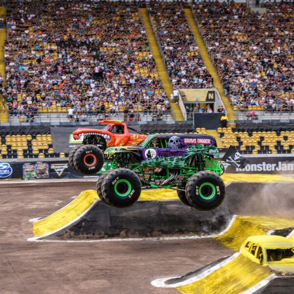 Foto: © 2023 Michael Schmid Ein dynamisches Bild, das einen Monster Jam-Wettbewerb im Rudolf-Harbig-Stadion in Dresden zeigt. Im Vordergrund springt der grüne Monster-Truck "Grave Digger" mit lila und schwarzen Flammen über eine gelbe Rampe, während er sich in der Luft befindet. Ein roter Monster-Truck ist direkt hinter ihm zu sehen, ebenfalls in der Luft über einer Rampe. Unter den Rampen sind die Überreste von zwei gelben Autos zu erkennen. Das Feld ist mit brauner Erde bedeckt, und im Hintergrund sind die gelben Tribünen des Stadions mit zahlreichen Zuschauern gefüllt. Das Wetter ist hell und sonnig, was auf eine Tagesveranstaltung hindeutet.
