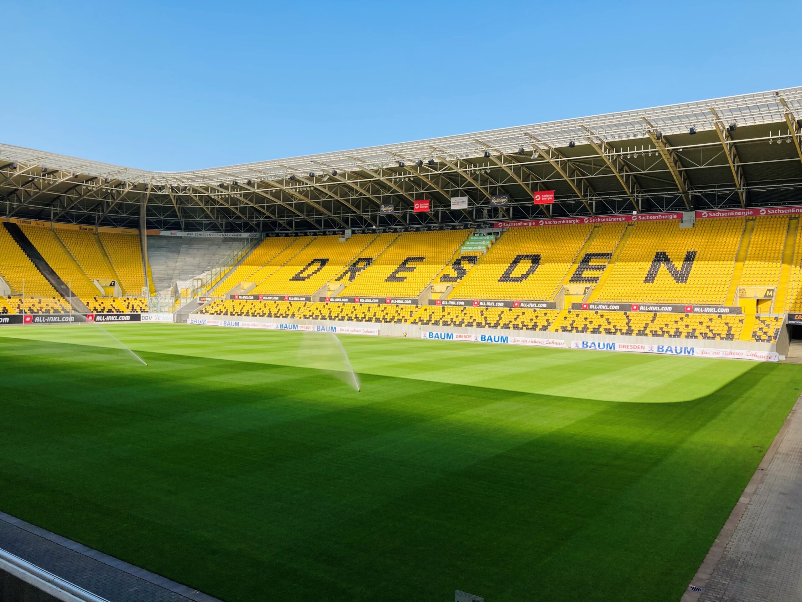 Ein Panoramabild des Rudolf-Harbig-Stadions in Dresden, aufgenommen an einem sonnigen Tag. Das Stadion ist leer, der grüne Rasen des Spielfelds ist frisch gemäht und wird bewässert. Die Sitzreihen sind in Gelb und Schwarz gehalten, mit dem Wort „DRESDEN“ in Schwarz auf den gelben Sitzen geschrieben. Der Himmel ist klar und blau.