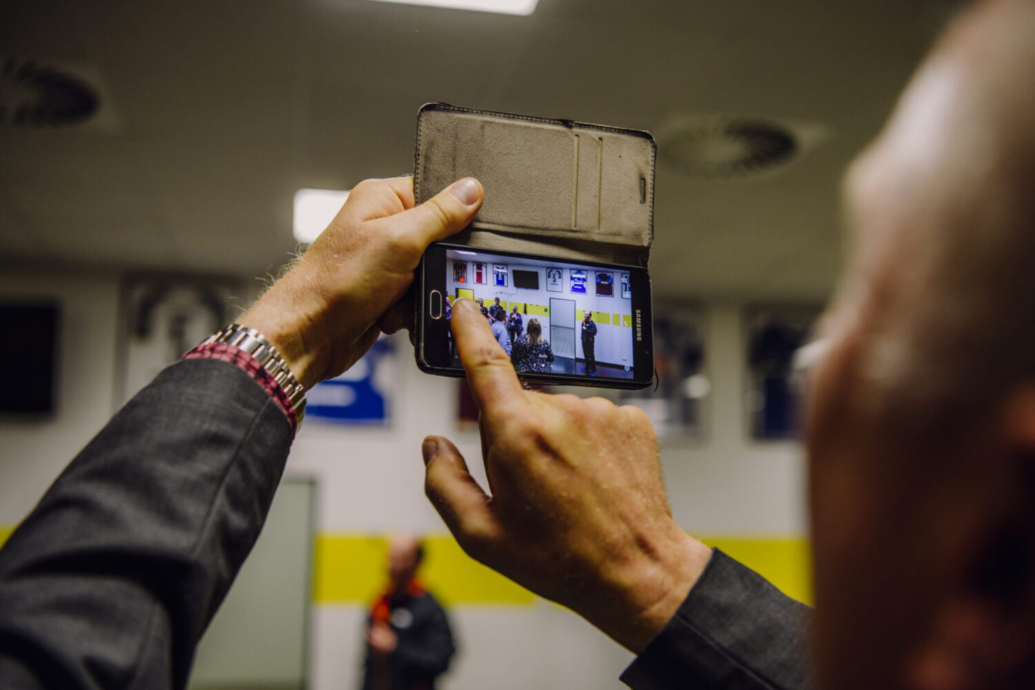 Aus der Ich-Perspektive eines Mannes, der ein Smartphone mit einer aufklappbaren Hülle in der Hand hält und mit dem Finger den Bildschirm berührt. Auf dem Smartphone-Bildschirm ist eine Szene mit mehreren Personen in einem Raum zu sehen, wahrscheinlich eine Aufnahme oder ein Live-Stream. Der Mann trägt ein Hemd oder Jackett und eine Armbanduhr mit einem pinken Band. Im unscharfen Hintergrund sind gelbe und weiße Wände, Leuchtstoffröhren an der Decke und andere Personen zu erkennen.
