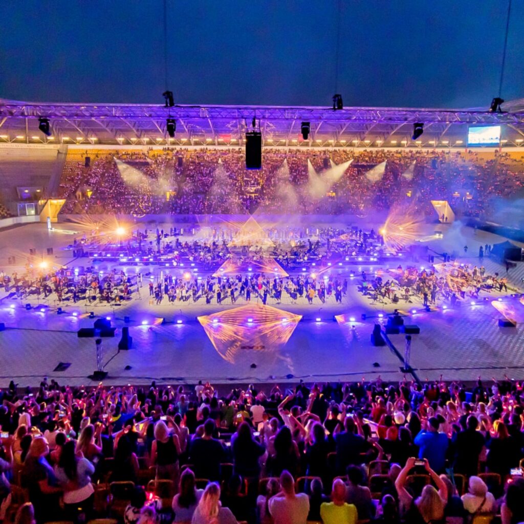 Dieses Bild zeigt ein Nachtkonzert in einem großen, beleuchteten Stadion. Die Bühne in der Mitte des Feldes ist hell erleuchtet mit blauen, violetten und orangefarbenen Lichtern, die eine große Menschenmenge beleuchten, die zu Füßen der Bühne versammelt ist. Das Stadion ist mit Tausenden von Zuschauern gefüllt, die die Aufführung genießen. Der Himmel über dem Stadion ist dunkel, was die lebhaften Lichter des Konzerts noch mehr hervorhebt.