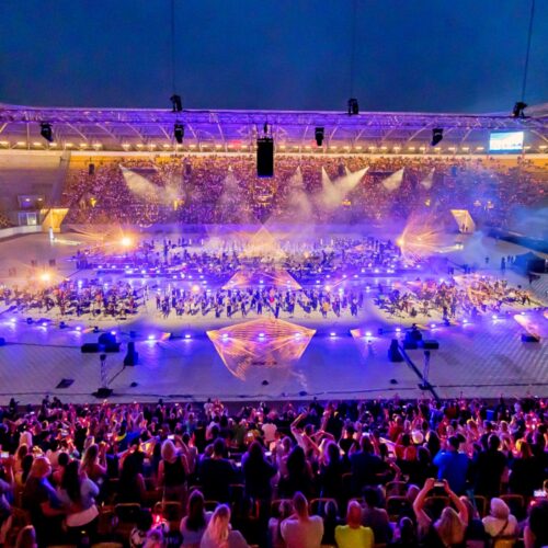 Dieses Bild zeigt ein Nachtkonzert in einem großen, beleuchteten Stadion. Die Bühne in der Mitte des Feldes ist hell erleuchtet mit blauen, violetten und orangefarbenen Lichtern, die eine große Menschenmenge beleuchten, die zu Füßen der Bühne versammelt ist. Das Stadion ist mit Tausenden von Zuschauern gefüllt, die die Aufführung genießen. Der Himmel über dem Stadion ist dunkel, was die lebhaften Lichter des Konzerts noch mehr hervorhebt.