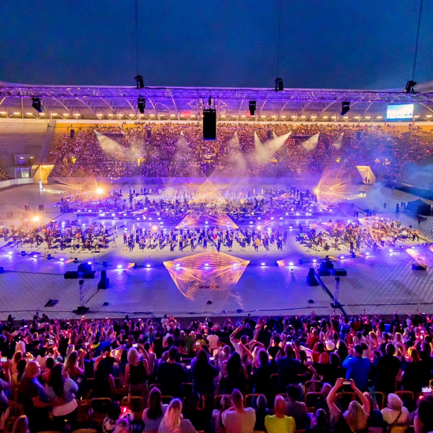 Ein weites Bild einer Stadionveranstaltung bei Nacht. Ein großer Teil des Bildes wird von der großen, hell beleuchteten Bühne im Zentrum eingenommen. Die Bühne ist von Tausenden von Menschen überfüllt. Einige tanzen, andere führen Choreografien auf. Viele der Darsteller tragen leuchtende Kostüme in verschiedenen Farben und halten leuchtende Gegenstände in den Händen. Die Tribünen um die Bühne herum sind voll mit Zuschauern. Der Himmel über dem Stadion ist dunkel und klar.