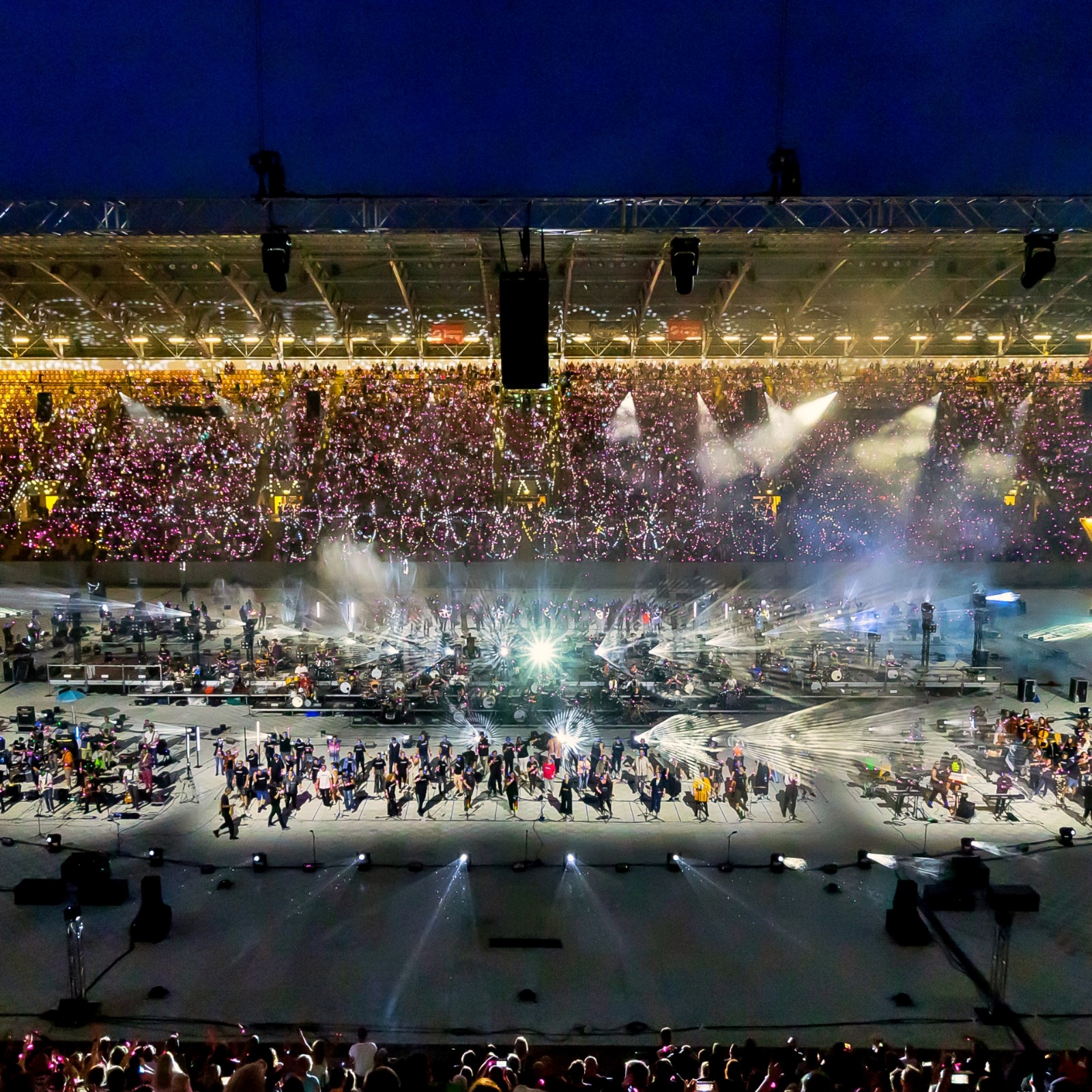 Ein hochwinkeliges Luftbild zeigt eine Menschenmenge, die ein Sportstadion füllt, dessen Tribünen bis zum Rand mit Zuschauern gefüllt sind. Die Tribünen leuchten in einem warmen, goldenen Licht. Der Boden des Stadions ist dunkel und scheint aus der Ferne von großen Lichtern von oben beleuchtet zu werden, die helle, konische Strahlen erzeugen. Auf dem Boden tanzt eine große Menschenmenge. Ihr Licht und ihre Bewegung schaffen ein Spektakel, das einen starken Kontrast zu den sitzenden Menschen auf den Tribünen bildet.