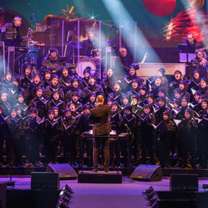 © 2024 Michael Schmidt Ein Chor, bestehend aus vielen Sängern in dunklen Mänteln, steht auf einer Bühne und singt. Im Vordergrund steht ein Dirigent von hinten, der den Chor leitet. Die Bühne ist von oben mit weißen Lichtstrahlen beleuchtet, die durch die Luft schneiden und eine festliche Atmosphäre erzeugen. Im Hintergrund sind Musiker und weitere Bühnenelemente zu erkennen, die leicht unscharf sind.