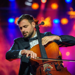© 2024 Michael Schmidt Ein Mann mit dunklem Haar und Bart, bekleidet mit einem Smoking und Fliege, spielt ein Cello auf der Bühne. Sein Blick ist auf das Instrument gerichtet. Der Hintergrund ist unscharf mit warmen, bunten Lichtern in Rot-, Orange- und Blautönen.