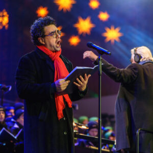 © 2024 Michael Schmidt Ein Mann mit lockigem dunklem Haar, einer Brille und einem roten Schal singt mit offenem Mund in ein Mikrofon und hält Notenblätter in seinen Händen. Er trägt einen dunklen Mantel. Rechts neben ihm steht ein weiterer Mann mit weißem Haar und Kopfhörern, der ebenfalls einen dunklen Mantel trägt und mit ausgestrecktem Arm dirigiert. Im Hintergrund sind unscharfe goldene Sternenlichter und blaue Lichter zu sehen. Unten links im Bild ist ein Teil eines Chores zu sehen.