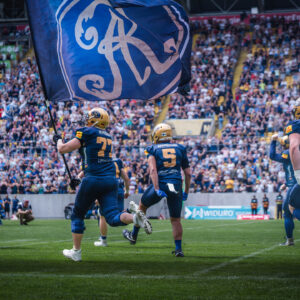 © LeoZiems Auf dem Bild sind American-Football-Spieler in dunklen Uniformen mit gelben Akzenten und Helmen zu sehen, die auf einem grünen Spielfeld in Richtung einer voll besetzten Tribüne laufen. Einer der Spieler trägt eine große blaue Fahne mit einem goldenen Emblem. Die Zuschauerränge im Hintergrund sind voll mit Fans.