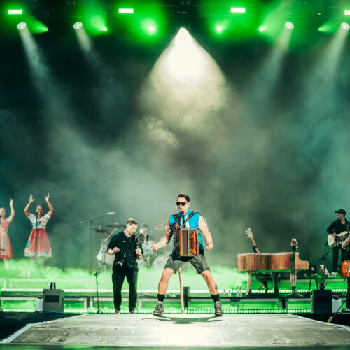 "Der Sänger Andreas Gabalier steht auf einer hell beleuchteten Bühne mit einem Akkordeon, umgeben von seiner Band und Effekten wie Nebel, während grünes Licht auf die Bühne fällt.