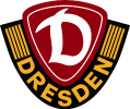 Das Logo von Dynamo Dresden, einem Fußballverein. Es zeigt ein dunkelrotes Schild mit einem stilisierten weißen "D" in der Mitte, das für Dresden steht. Das Schild ist von einem schwarzen, schildförmigen Rand umgeben, auf dem in Großbuchstaben "DRESDEN" in Gold zu lesen ist. Links und rechts des roten Schildes, innerhalb des schwarzen Rahmens, sind goldene vertikale Streifen abgebildet. Der Hintergrund des Bildes ist transparent oder als graues und weißes Schachbrettmuster dargestellt.