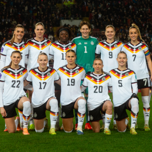 Eine Fußballmannschaft posiert für ein Gruppenfoto auf dem Spielfeld eines großen Stadions. Die Spieler tragen weiße Trikots mit grünen und orangefarbenen Akzenten; einige stehen, andere knien in der ersten Reihe. Im Hintergrund sind die beleuchteten Tribünen und weitere Personen am Spielfeldrand zu sehen.