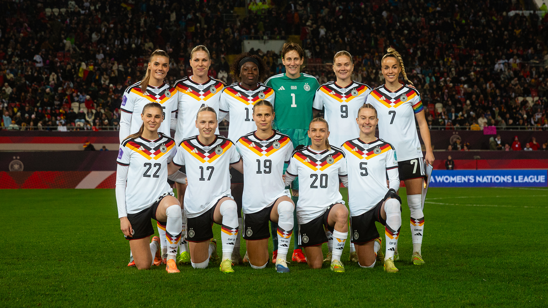 Eine Fußballmannschaft posiert für ein Gruppenfoto auf dem Spielfeld eines großen Stadions. Die Spieler tragen weiße Trikots mit grünen und orangefarbenen Akzenten; einige stehen, andere knien in der ersten Reihe. Im Hintergrund sind die beleuchteten Tribünen und weitere Personen am Spielfeldrand zu sehen.