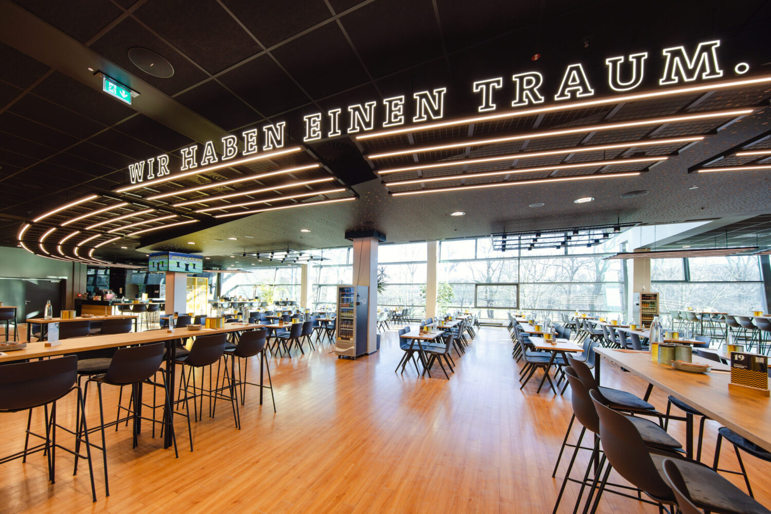 ©Claudia Jacquemin/Stadion Dresden Projektgesellschaft mbH & Co. KG Ein heller, moderner Raum mit Holzboden und einer großen Fensterfront im Hintergrund. An der dunklen Decke befindet sich eine geschwungene Lichtinstallation mit dem leuchtenden Schriftzug „WIR HABEN EINEN TRAUM.“. Im Vordergrund stehen lange Holztische mit dunklen Barhockern.