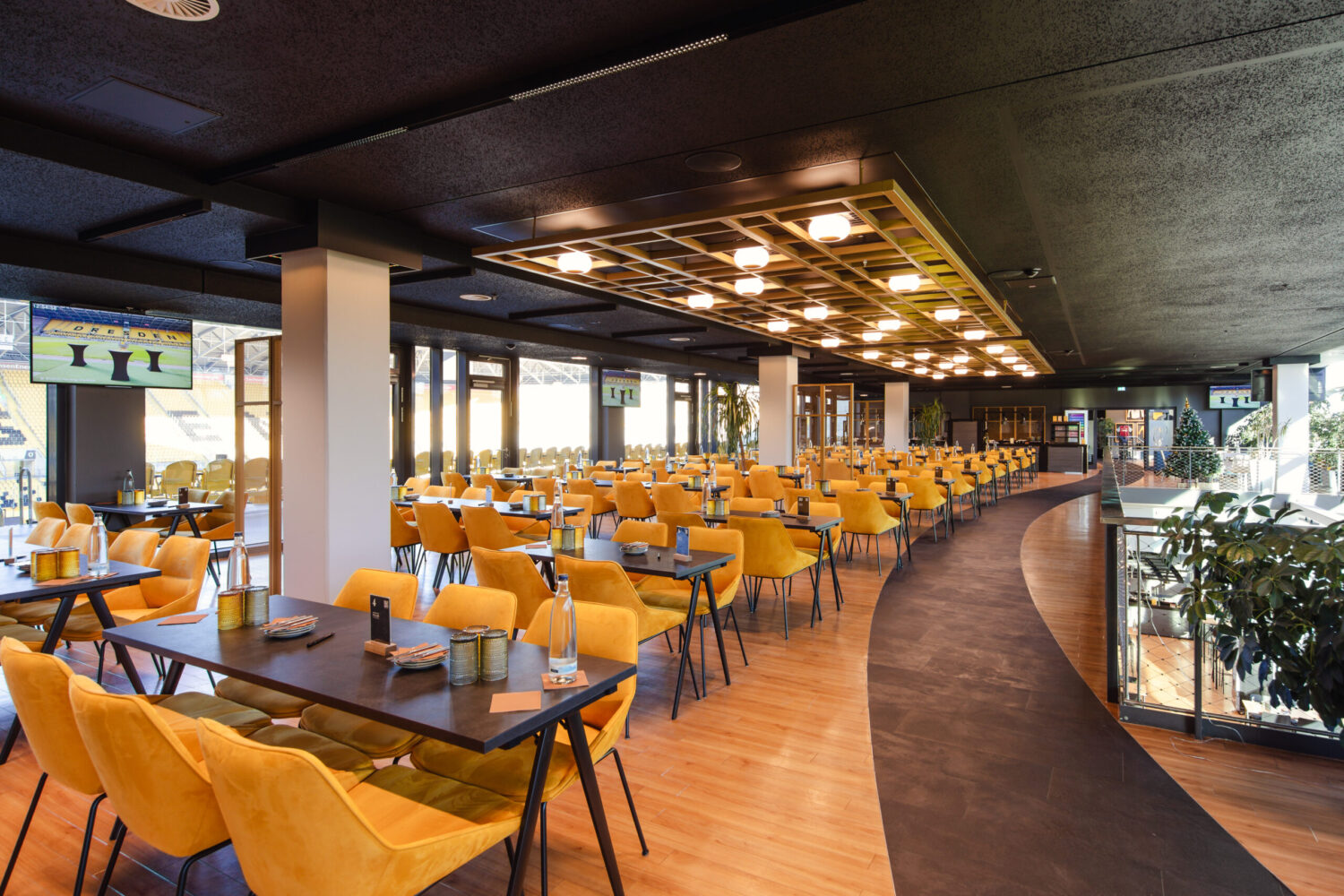 ©Claudia Jacquemin/Stadion Dresden Projektgesellschaft mbH & Co. KG Ein moderner, weitläufiger Restaurant- oder Loungebereich mit zahlreichen schwarzen Tischen und leuchtend gelben Polsterstühlen. Der Raum hat eine dunkle Decke mit einem auffälligen, beleuchteten Gitterelement in der Mitte und eine große Fensterfront, die viel Tageslicht hereinlässt.