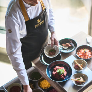 ©Supreme Sports Hospitality GmbH Ein Koch in weißer Uniform und schwarzer Schürze mit goldenem Logo bereitet an einer Live-Cooking-Station Speisen zu. Er richtet kleine Schalen mit verschiedenen Zutaten und Toppings an, die in einer übersichtlichen Auslage vor ihm präsentiert werden. Die Perspektive ist leicht von oben fotografiert.