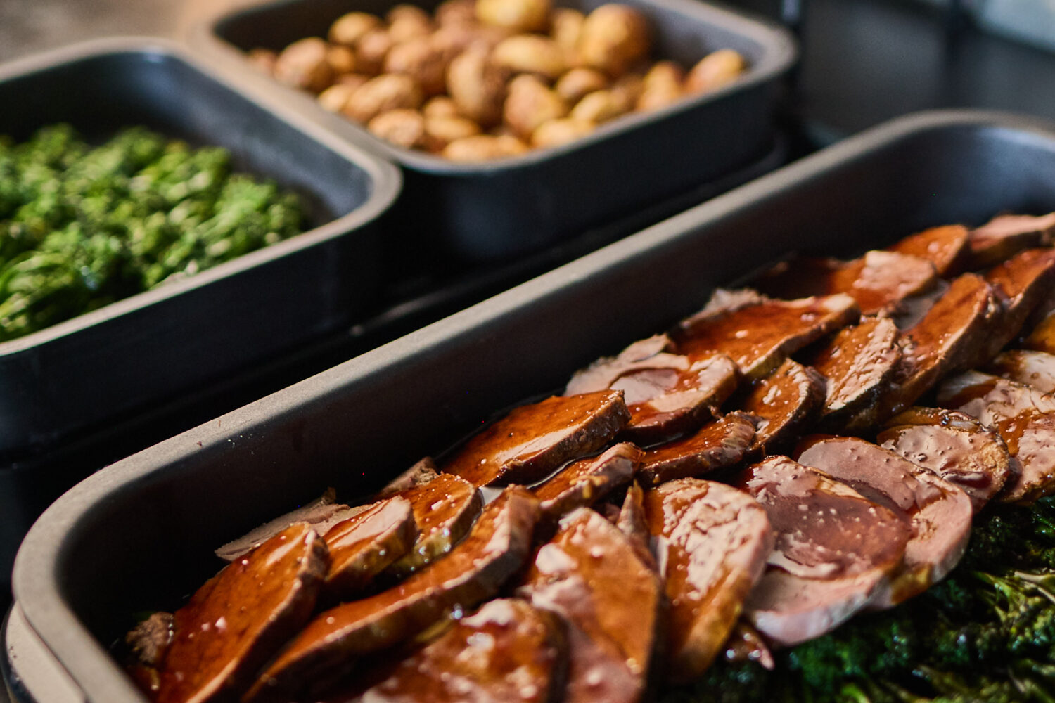 ©Supreme Sports Hospitality GmbH Nahaufnahme einer Buffet-Station mit warmen Speisen. Im Vordergrund steht eine Servierschale mit zart geschnittenem, glasiertem Braten auf einem Bett aus grünem Gemüse. Dahinter befinden sich weitere Schalen mit Beilagen wie Röstkartoffeln und Brokkoli sowie ordentlich gestapeltes Geschirr.