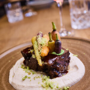 ©Supreme Sports Hospitality GmbH Nahaufnahme eines ästhetisch angerichteten Hauptgangs auf einem rustikalen braunen Teller. Ein Stück geschmorter Braten mit glänzender Sauce ruht auf einem cremigen Püreespiegel, garniert mit bunten Mini-Karotten und Kräutern. Im unscharfen Hintergrund ist ein Weinglas mit Rotwein auf einem Holztisch zu erkennen.