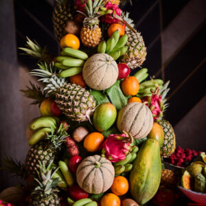 ©Supreme Sports Hospitality GmbH Anfrage erfolgreich Gemini said Ein üppiges, pyramidenförmiges Arrangement aus verschiedenen exotischen und heimischen Früchten auf einem Buffet. Zu sehen sind unter anderem mehrere ganze Ananas, Cantaloupe-Melonen, Drachenfrüchte, Papayas, Mangos, Kiwis sowie Bananenstauden und Zitrusfrüchte. Im Vordergrund stehen kleine Teller mit mundgerecht geschnittenen Fruchtstücken. Das Ganze ist stimmungsvoll vor einem dunklen Hintergrund mit grafischem Muster angerichtet.