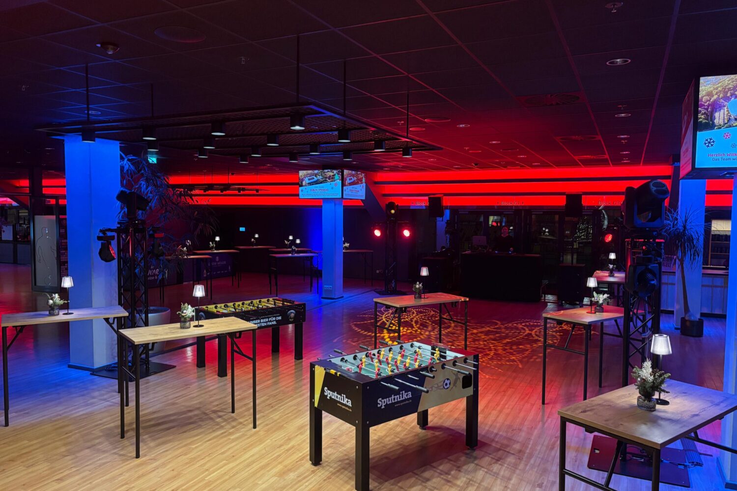 Ein stimmungsvoll beleuchteter Veranstaltungsraum bei Nacht. Im Vordergrund stehen mehrere Tischkicker-Geräte und Stehtische mit kleinen Lampen auf einem hellen Holzboden. Der Raum wird durch markante rote Lichtleisten an der Rückwand und blau leuchtende Säulen in ein modernes, farbenfrohes Licht getaucht.