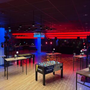 Ein stimmungsvoll beleuchteter Veranstaltungsraum bei Nacht. Im Vordergrund stehen mehrere Tischkicker-Geräte und Stehtische mit kleinen Lampen auf einem hellen Holzboden. Der Raum wird durch markante rote Lichtleisten an der Rückwand und blau leuchtende Säulen in ein modernes, farbenfrohes Licht getaucht.