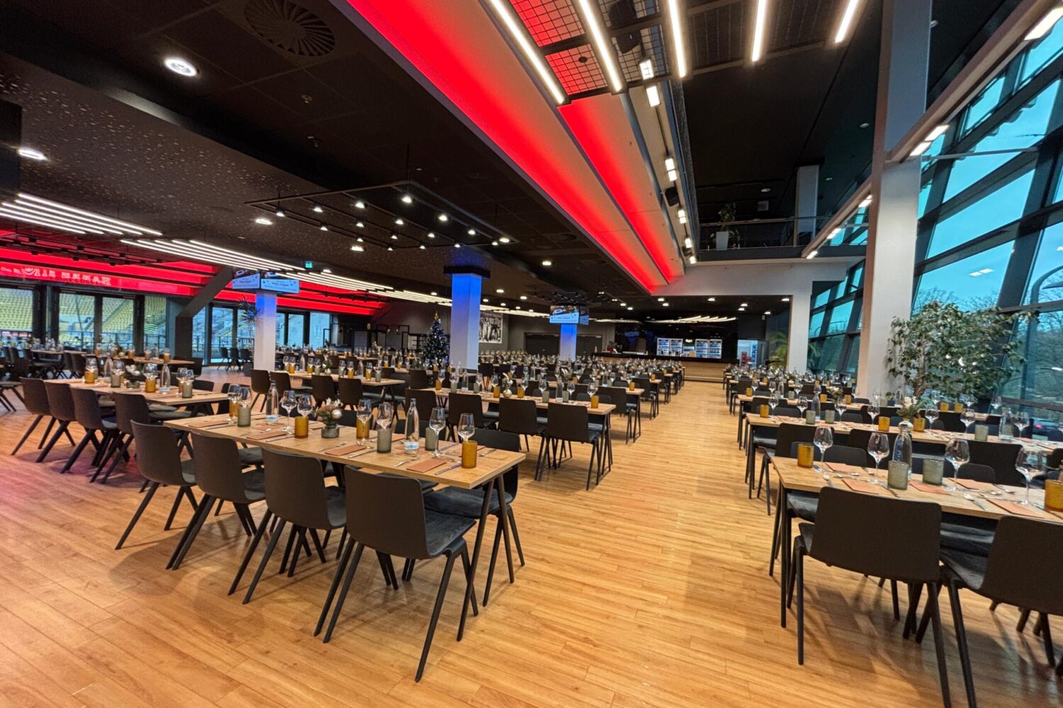 Ein weitläufiger, modern gestalteter Veranstaltungssaal mit langen Holztiischen und dunklen Stühlen, die in ordentlichen Reihen aufgestellt sind. Die Decke ist mit markanten roten und weißen Lichtbändern sowie runden Strahlern versehen; blaue Akzentbeleuchtung an den Säulen sorgt für eine dynamische Atmosphäre. Große Fensterfronten auf der rechten Seite geben den Blick nach draußen frei.