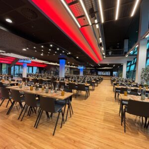 Ein weitläufiger, modern gestalteter Veranstaltungssaal mit langen Holztiischen und dunklen Stühlen, die in ordentlichen Reihen aufgestellt sind. Die Decke ist mit markanten roten und weißen Lichtbändern sowie runden Strahlern versehen; blaue Akzentbeleuchtung an den Säulen sorgt für eine dynamische Atmosphäre. Große Fensterfronten auf der rechten Seite geben den Blick nach draußen frei.