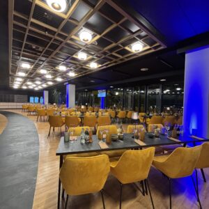 Ein eleganter Gastronomiebereich mit langen dunklen Tischen und auffälligen, senfgelben Polstersesseln. Die Decke ist mit einem quadratischen Holzraster und warmen Leuchten gestaltet. Blau beleuchtete Säulen und große Fensterfronten im Hintergrund verleihen dem Raum eine exklusive Abendatmosphäre.