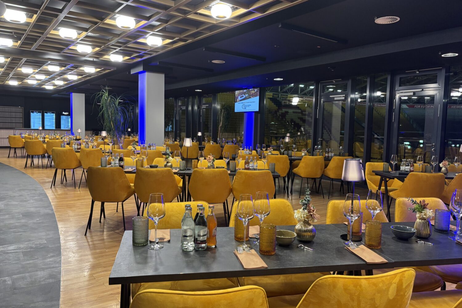 Eine stimmungsvolle Innenansicht eines modernen Eventbereichs bei Abendbeleuchtung. Im Vordergrund steht ein eingedeckter schwarzer Tisch mit Weingläsern und Wasserflaschen, umgeben von gepolsterten, senfgelben Stühlen. Die Decke ist durch ein markantes quadratisches Holzraster mit integrierten runden Leuchten charakterisiert, während blaue Lichtakzente an den Säulen und große Glasfronten im Hintergrund für ein elegantes Ambiente sorgen.